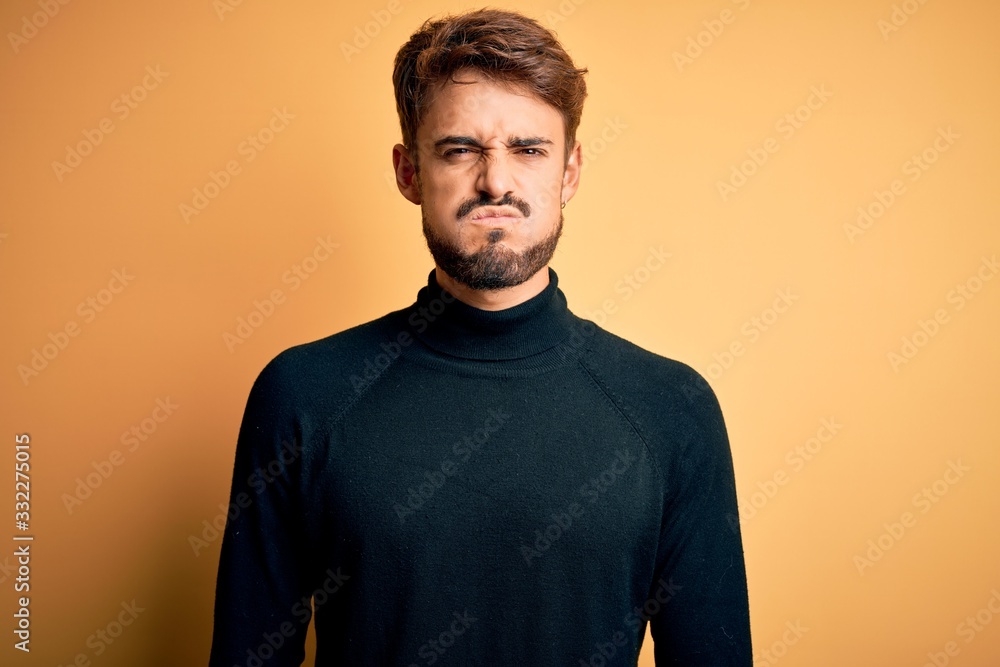 Young handsome man with beard wearing turtleneck sweater standing over yellow background puffing cheeks with funny face. Mouth inflated with air, crazy expression. Young handsome man with beard wearing turtleneck sweater standing over yellow background puffing cheeks with funny face. Mouth inflated with air, crazy expression.