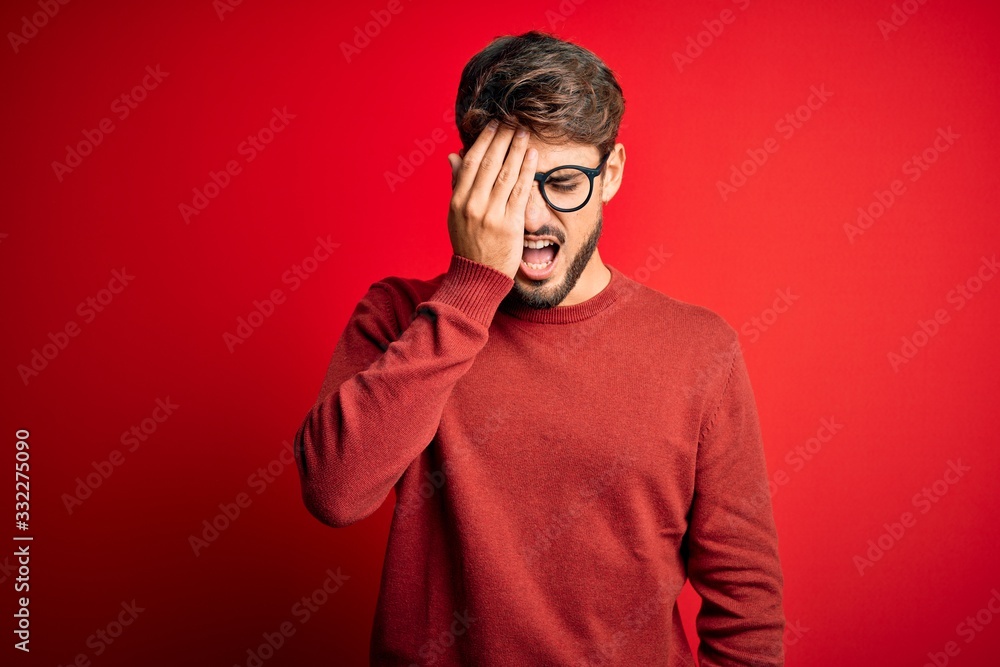 Young handsome man with beard wearing glasses and sweater standing over red background Yawning tired covering half face, eye and mouth with hand. Face hurts in pain.