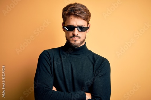 Young man wearing thug life fanny sunglasses standing over isolated yellow background skeptic and nervous, disapproving expression on face with crossed arms. Negative person.