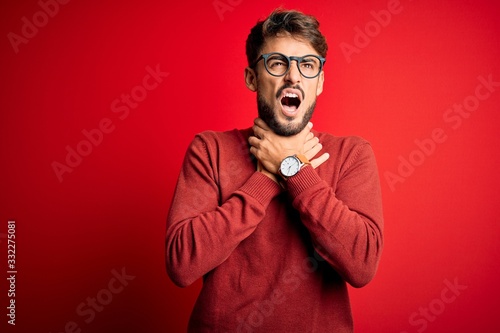 Young handsome man with beard wearing glasses and sweater standing over red background shouting suffocate because painful strangle. Health problem. Asphyxiate and suicide concept.