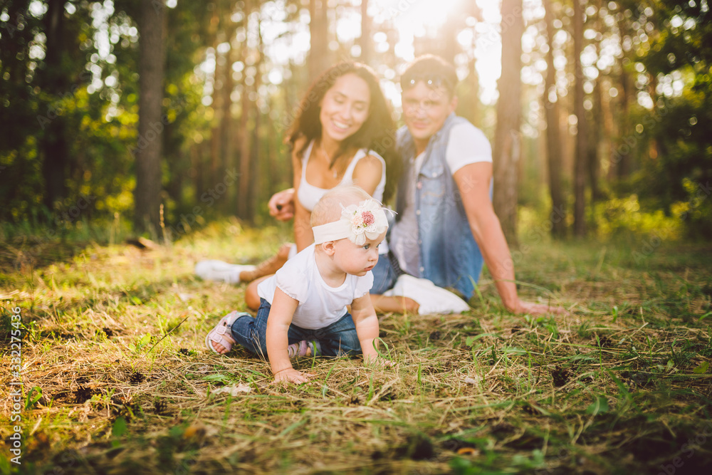 Fototapeta premium little girl one year on the background of parents resting lying on grass learning to walk on nature in the park. The first steps of a child in nature outside the city in the forest