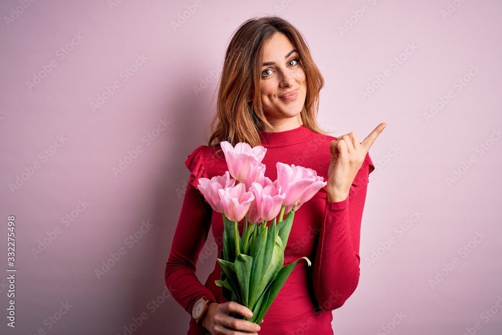Young beautiful brunette woman holding bouquet of pink tulips over isolated background cheerful with a smile on face pointing with hand and finger up to the side with happy and natural expression Young beautiful brunette woman holding bouquet of pink tulips over isolated background cheerful with a smile on face pointing with hand and finger up to the side with happy and natural expression