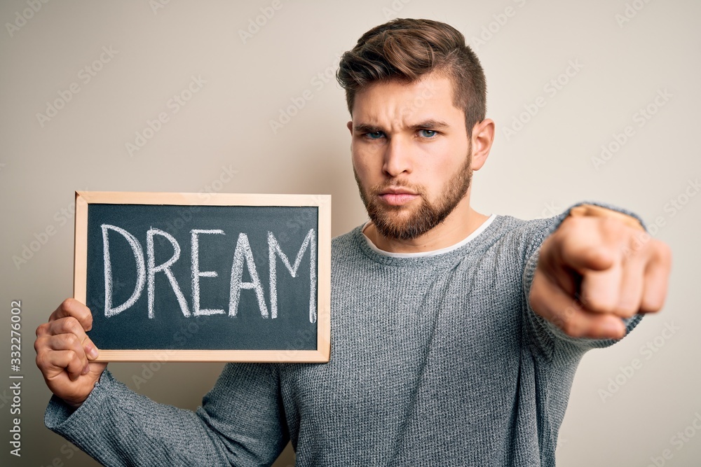 Young blond man with beard and blue eyes holding blackboard with dream message pointing with finger to the camera and to you, hand sign, positive and confident gesture from the front