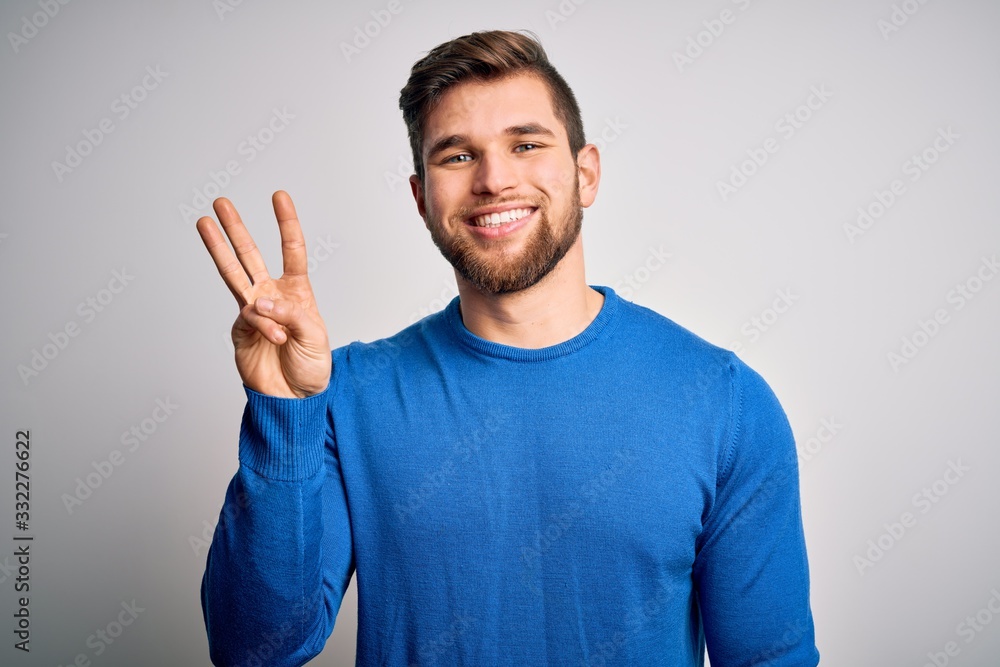 Young handsome blond man with beard and blue eyes wearing casual sweater showing and pointing up with fingers number three while smiling confident and happy. Young handsome blond man with beard and blue eyes wearing casual sweater showing and pointing up with fingers number three while smiling confident and happy.