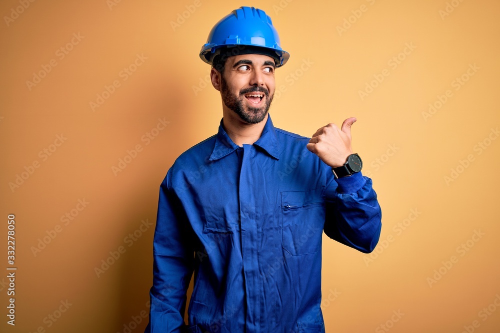 Mechanic man with beard wearing blue uniform and safety helmet over yellow background smiling with happy face looking and pointing to the side with thumb up.