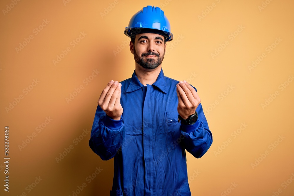 Mechanic man with beard wearing blue uniform and safety helmet over yellow background doing money gesture with hands, asking for salary payment, millionaire business