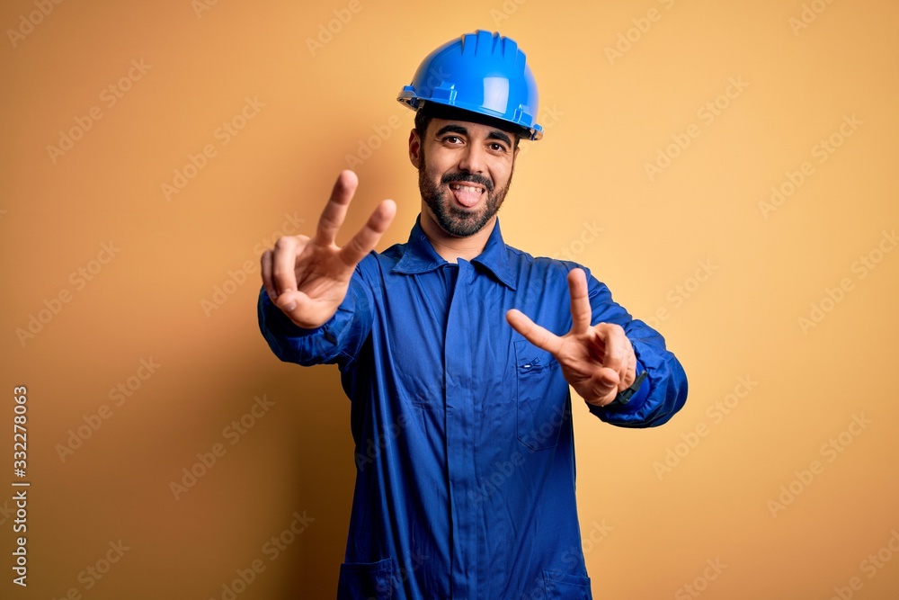 Mechanic man with beard wearing blue uniform and safety helmet over yellow background smiling with tongue out showing fingers of both hands doing victory sign. Number two.