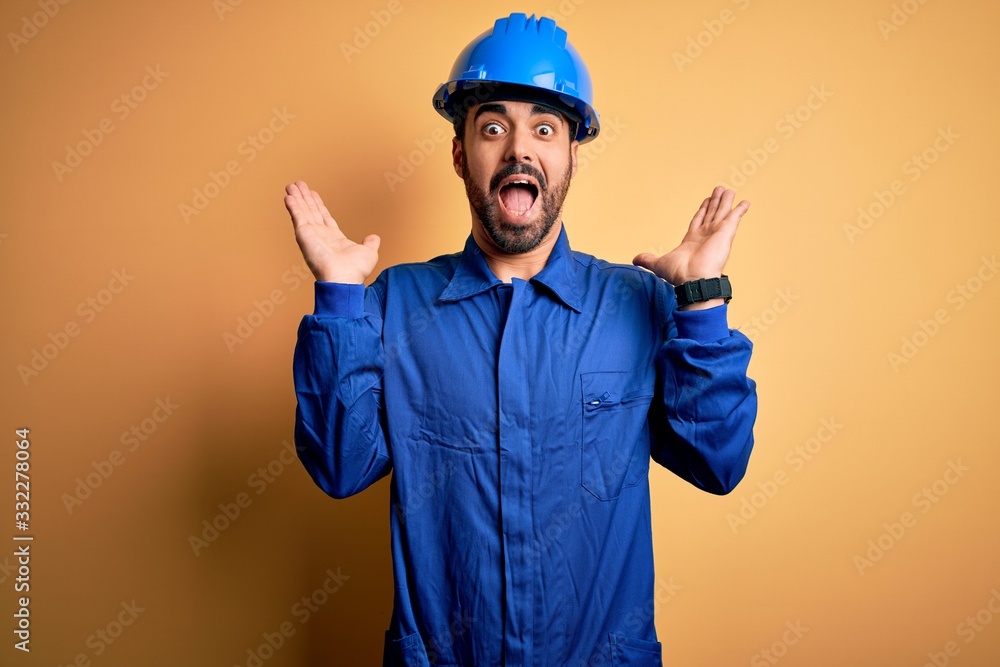 Mechanic man with beard wearing blue uniform and safety helmet over yellow background celebrating crazy and amazed for success with arms raised and open eyes screaming excited. Winner concept