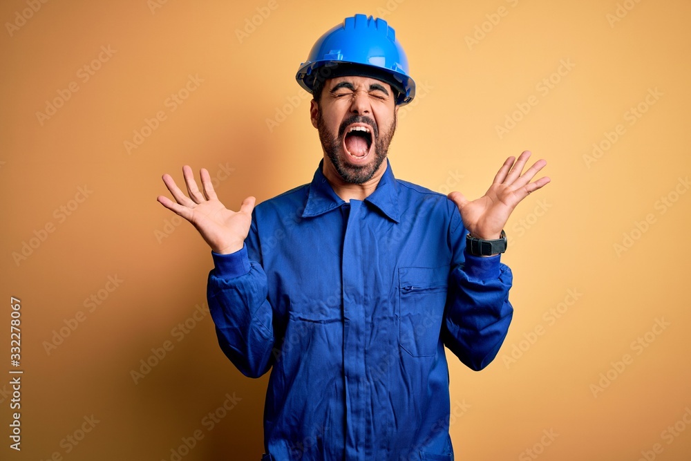 Mechanic man with beard wearing blue uniform and safety helmet over yellow background celebrating mad and crazy for success with arms raised and closed eyes screaming excited. Winner concept