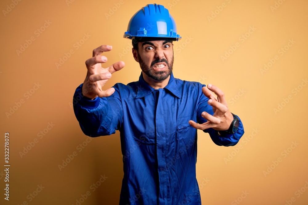 Mechanic man with beard wearing blue uniform and safety helmet over yellow background Shouting frustrated with rage, hands trying to strangle, yelling mad