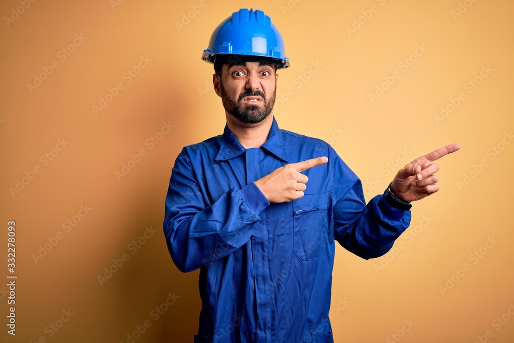 Mechanic man with beard wearing blue uniform and safety helmet over yellow background Pointing aside worried and nervous with both hands, concerned and surprised expression