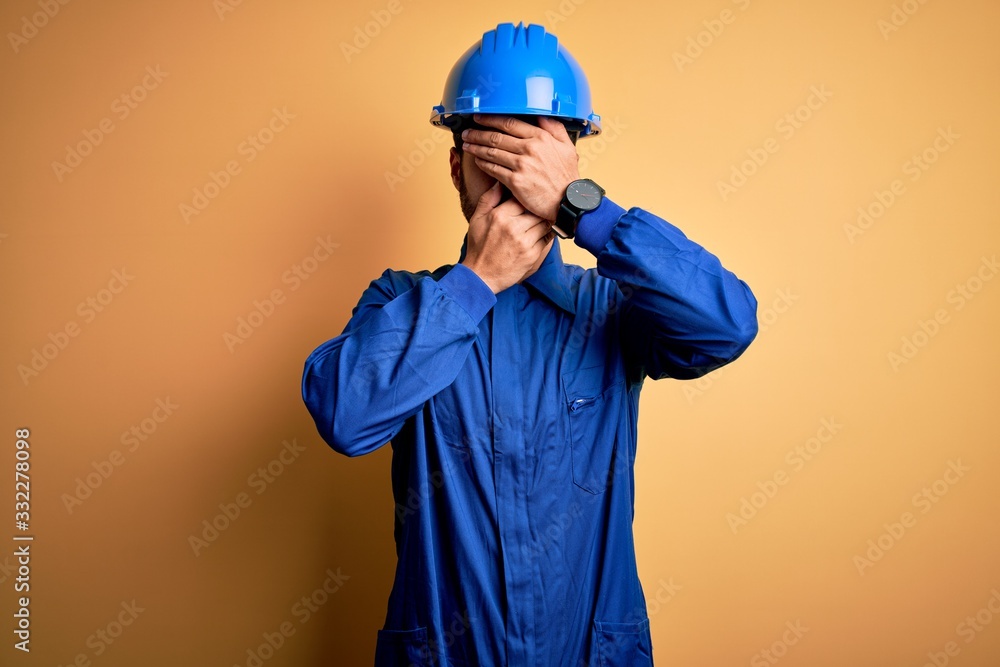 Mechanic man with beard wearing blue uniform and safety helmet over yellow background Covering eyes and mouth with hands, surprised and shocked. Hiding emotion