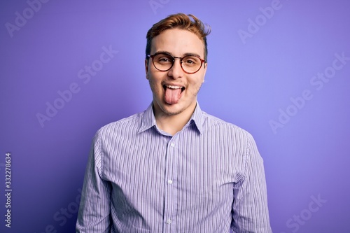 Young handsome redhead man wearing casual shirt and glasses over purple background sticking tongue out happy with funny expression. Emotion concept. © Krakenimages.com