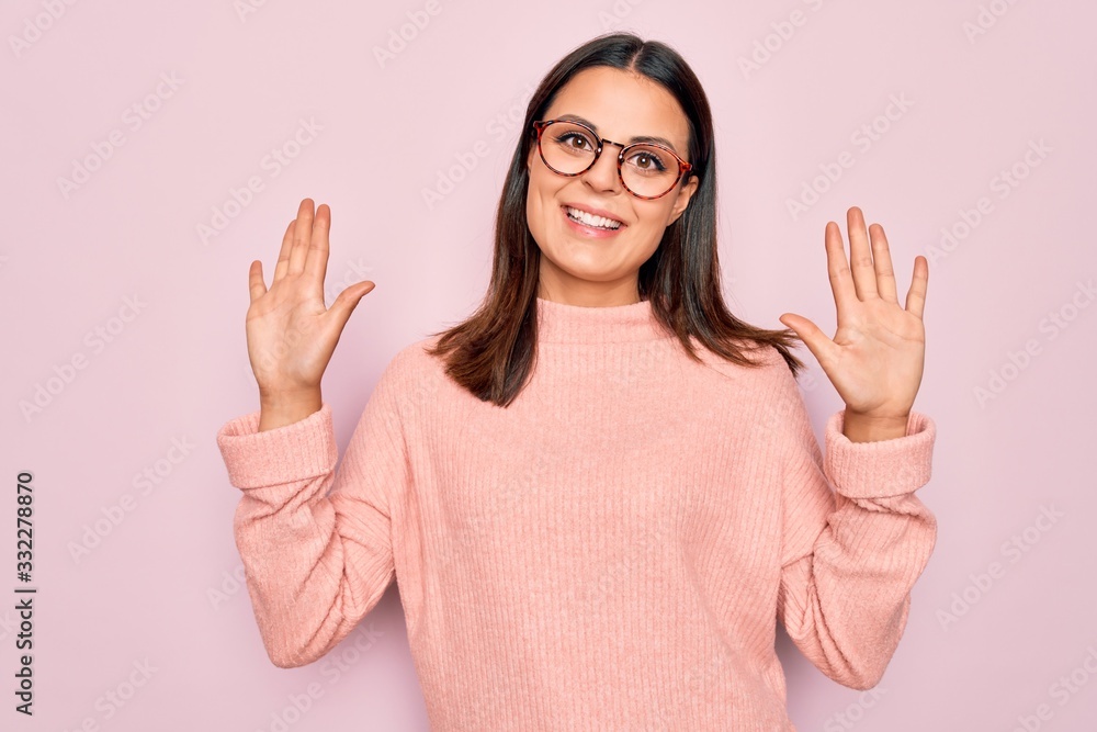 Young beautiful brunette woman wearing casual sweater and glasses over pink background showing and pointing up with fingers number ten while smiling confident and happy. Young beautiful brunette woman wearing casual sweater and glasses over pink background showing and pointing up with fingers number ten while smiling confident and happy.