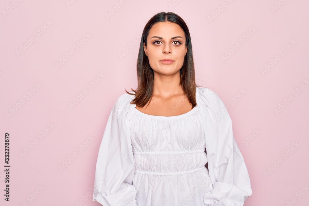 Fototapeta premium Young beautiful brunette woman wearing casual white dress standing over pink background with serious expression on face. Simple and natural looking at the camera.