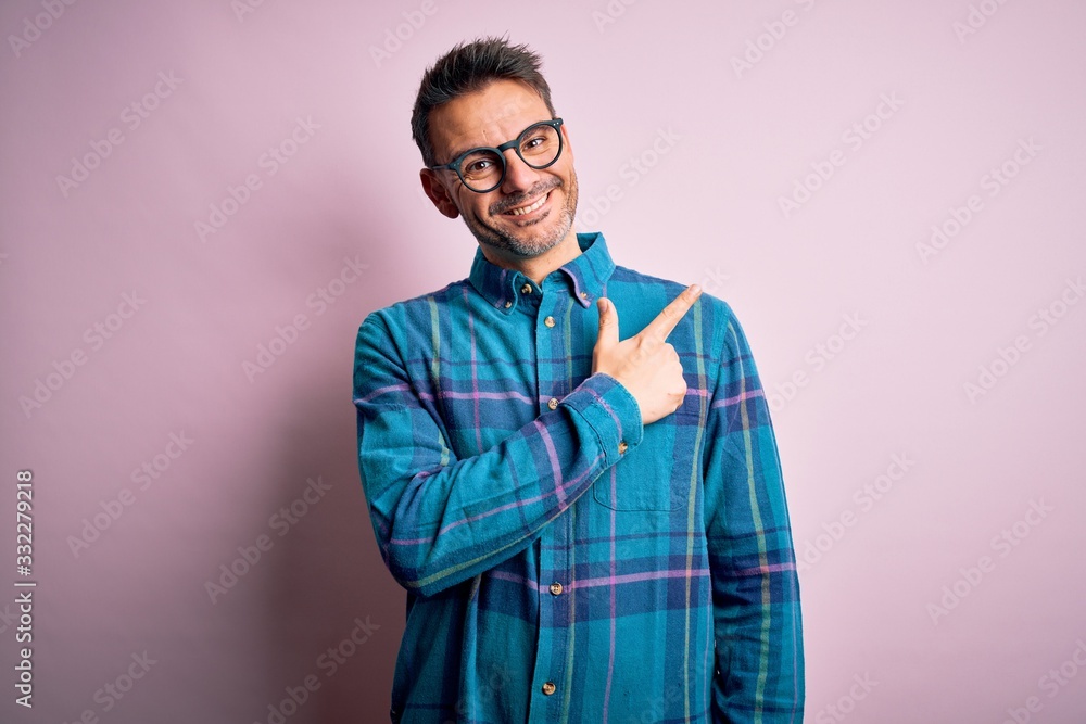Young handsome man wearing casual shirt and glasses standing over isolated pink background cheerful with a smile of face pointing with hand and finger up to the side with happy and natural expression Young handsome man wearing casual shirt and glasses standing over isolated pink background cheerful with a smile of face pointing with hand and finger up to the side with happy and natural expression