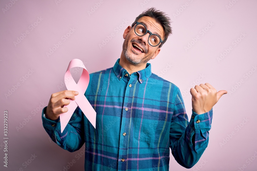 Young handsome man holding pink cancer ribbon symbol over isolated background pointing and showing with thumb up to the side with happy face smiling Young handsome man holding pink cancer ribbon symbol over isolated background pointing and showing with thumb up to the side with happy face smiling