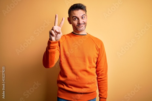 Young handsome man wearing orange casual sweater standing over isolated yellow background showing and pointing up with fingers number two while smiling confident and happy.