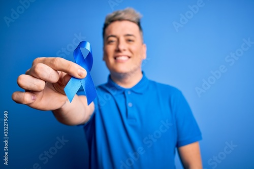 Close up of young man holding colon cancer awareness blue ribbon over isolated background with a happy face standing and smiling with a confident smile showing teeth