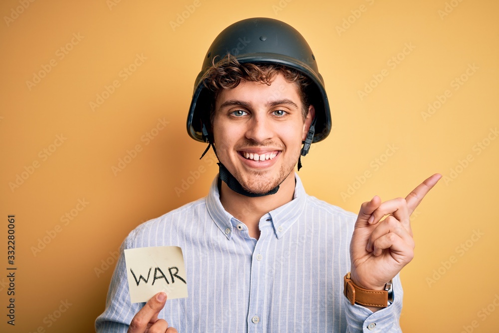 Young blond businessman with curly hair wearing helmet holding paper with war message very happy pointing with hand and finger to the side