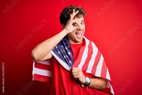 Young blond patriotic man with curly hair wearing USA flag celebrating independence day with happy face smiling doing ok sign with hand on eye looking through fingers
