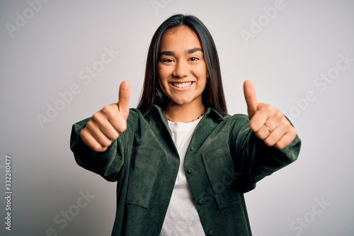 Young beautiful asian woman wearing casual shirt standing over isolated white background approving doing positive gesture with hand, thumbs up smiling and happy for success. Winner gesture.