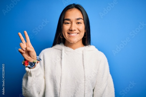 Young beautiful asian sportswoman wearing sweatshirt standing over isolated blue background smiling with happy face winking at the camera doing victory sign. Number two.