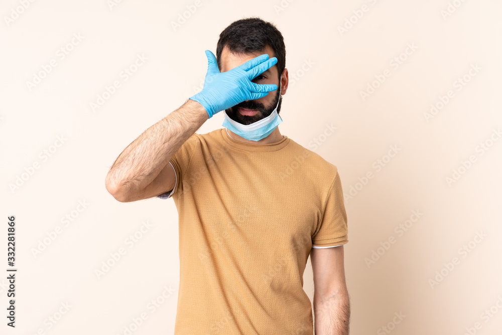 Caucasian man with beard protecting from the coronavirus with a mask and gloves over isolated background covering eyes by hands and smiling Caucasian man with beard protecting from the coronavirus with a mask and gloves over isolated background covering eyes by hands and smiling