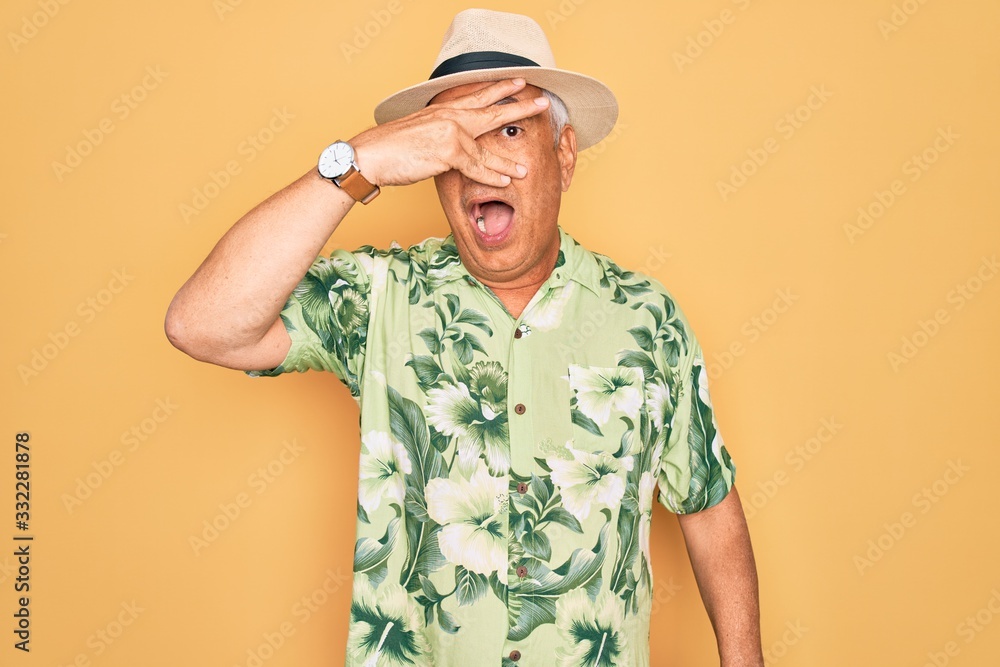 Middle age senior grey-haired man wearing summer hat and floral shirt on beach vacation peeking in shock covering face and eyes with hand, looking through fingers with embarrassed expression. Middle age senior grey-haired man wearing summer hat and floral shirt on beach vacation peeking in shock covering face and eyes with hand, looking through fingers with embarrassed expression.