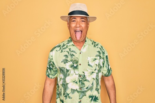 Middle age senior grey-haired man wearing summer hat and floral shirt on beach vacation sticking tongue out happy with funny expression. Emotion concept.