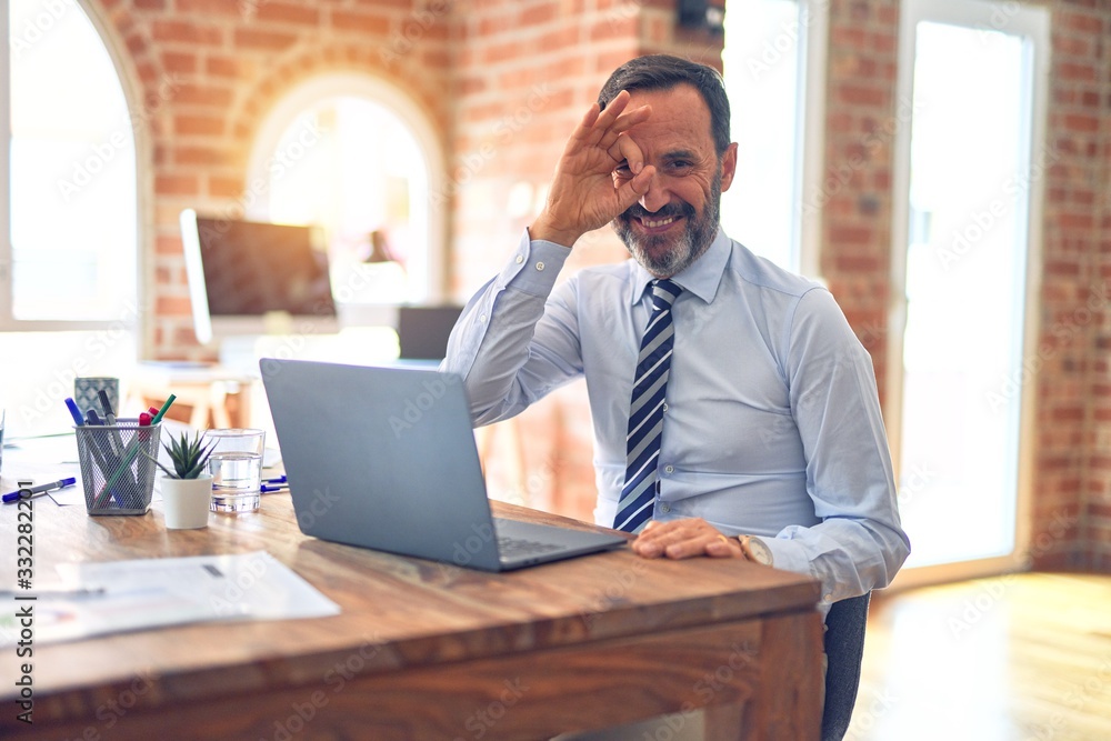Middle age handsome businessman wearing tie sitting using laptop at the office doing ok gesture with hand smiling, eye looking through fingers with happy face. Middle age handsome businessman wearing tie sitting using laptop at the office doing ok gesture with hand smiling, eye looking through fingers with happy face.