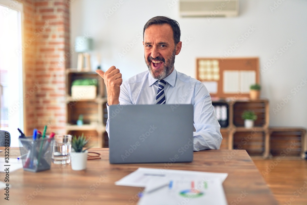 Middle age handsome businessman wearing tie sitting using laptop at the office smiling with happy face looking and pointing to the side with thumb up.
