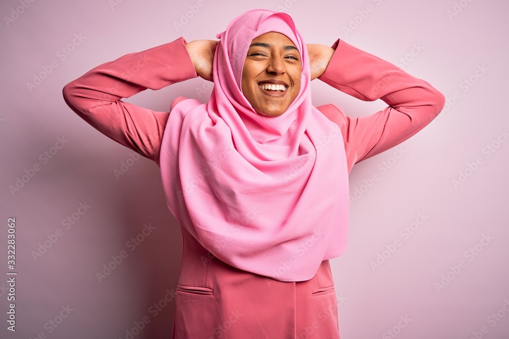 Fototapeta premium Young African American afro woman wearing muslim hijab over isolated pink background relaxing and stretching, arms and hands behind head and neck smiling happy