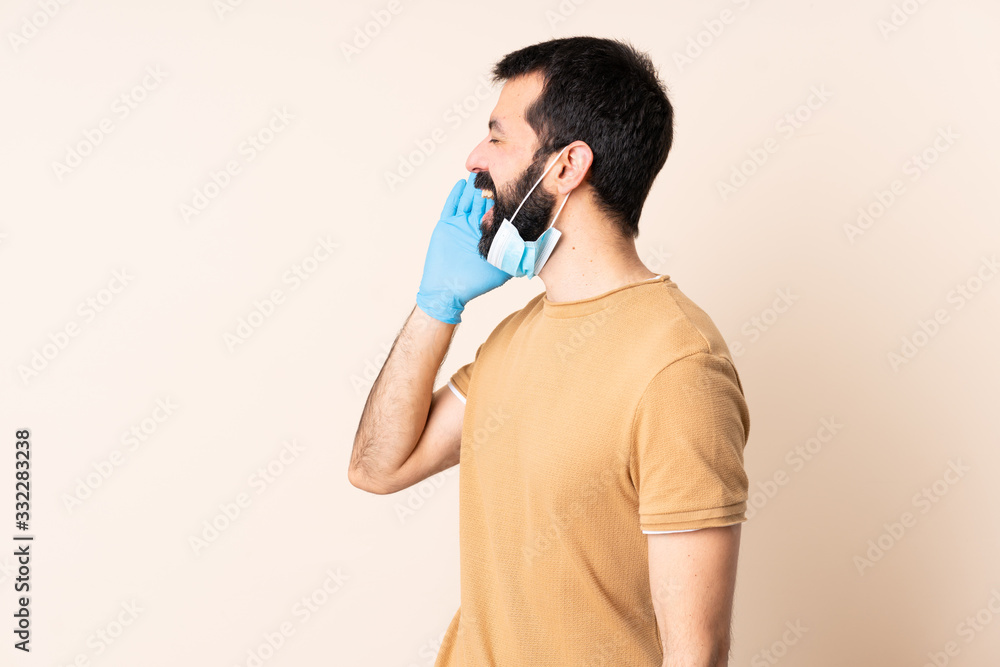 Caucasian man with beard protecting from the coronavirus with a mask and gloves over isolated background shouting with mouth wide open to the side Caucasian man with beard protecting from the coronavirus with a mask and gloves over isolated background shouting with mouth wide open to the side