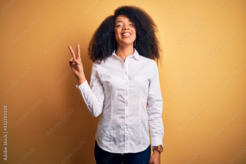 Young beautiful african american elegant woman with afro hair standing over yellow background smiling looking to the camera showing fingers doing victory sign. Number two. Young beautiful african american elegant woman with afro hair standing over yellow background smiling looking to the camera showing fingers doing victory sign. Number two.