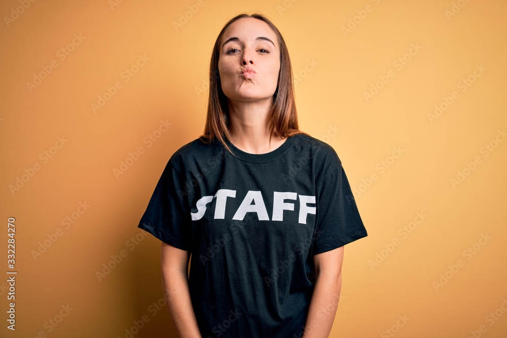 Young beautiful worker woman wearing staff uniform t-shirt over isolated yellow background looking at the camera blowing a kiss on air being lovely and sexy. Love expression. Young beautiful worker woman wearing staff uniform t-shirt over isolated yellow background looking at the camera blowing a kiss on air being lovely and sexy. Love expression.