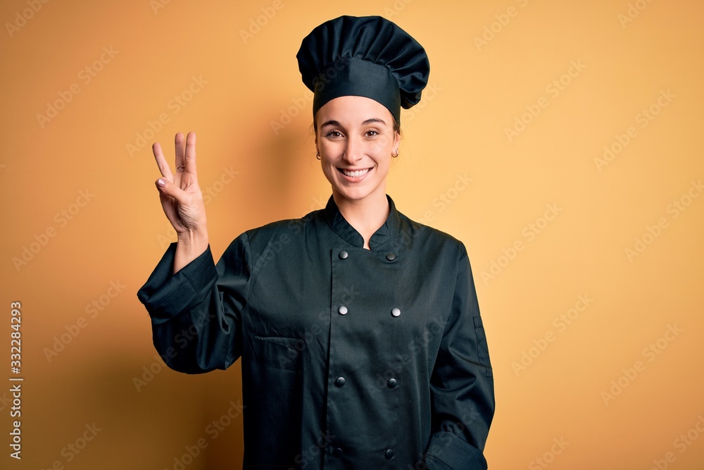 Young beautiful chef woman wearing cooker uniform and hat standing over yellow background showing and pointing up with fingers number three while smiling confident and happy. Young beautiful chef woman wearing cooker uniform and hat standing over yellow background showing and pointing up with fingers number three while smiling confident and happy.