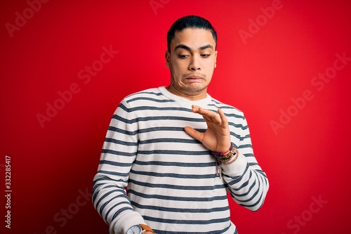 Young brazilian man wearing casual striped t-shirt standing over isolated red background disgusted expression, displeased and fearful doing disgust face because aversion reaction.