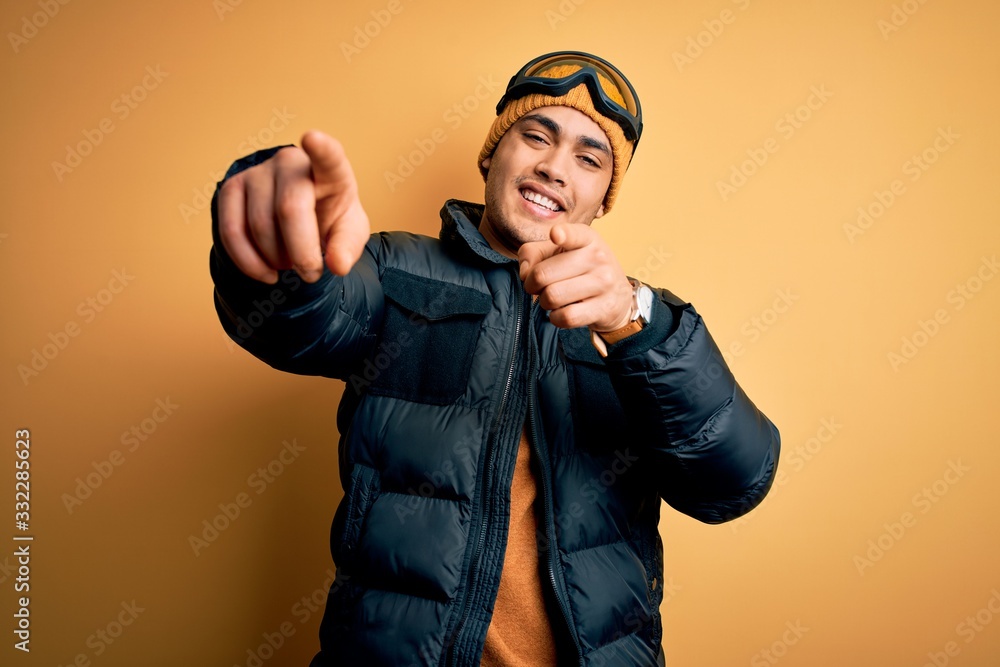 Young brazilian skier man wearing snow sportswear and ski goggles over yellow background pointing to you and the camera with fingers, smiling positive and cheerful Young brazilian skier man wearing snow sportswear and ski goggles over yellow background pointing to you and the camera with fingers, smiling positive and cheerful