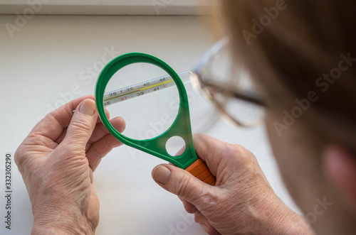 Elderly woman with glasses looks through round green magnifying glass holding in left hand mercury thermometer which indicating high temperature. Concept: Feverish heat harbinger of flu or coronavirus
