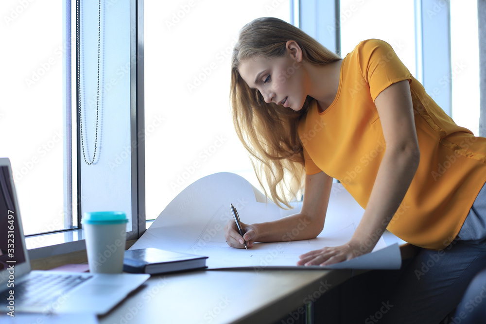 Beautiful modern woman is writing something down on the blueprint while working in the creative office Beautiful modern woman is writing something down on the blueprint while working in the creative office
