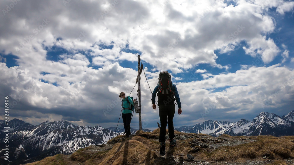 A couple with hiking backpacks standing on the top of Himmeleck in Austrian Alps. There is a massive mountain range in the back, partially covered with snow. Having fun, playtime. Cross on the top