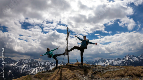 A couple with hiking backpacks standing on the top of Himmeleck in Austrian Alps. There is a massive mountain range in the back, partially covered with snow. Having fun, playtime. Cross on the top