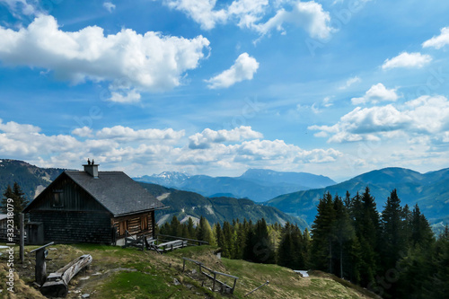 A small cottage located at the mountain ledge with the vast, panoramic view on the Alps in Austria. There are many mountain ranges in the back. Spring coming to the Alps. Dense forest