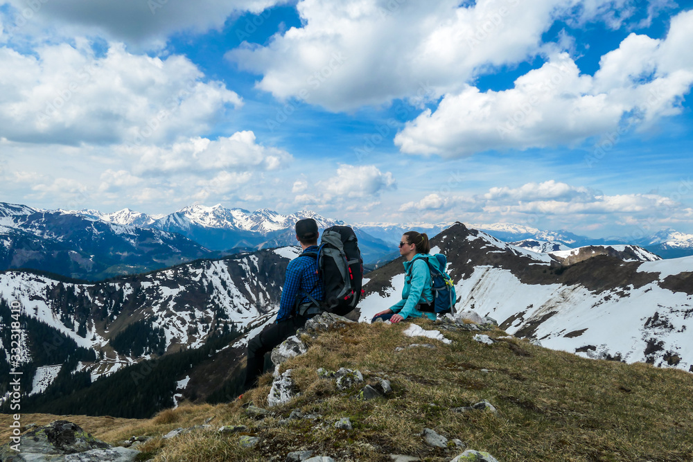Fototapeta premium Couple with big hiking backpacks resting on their way to Himmeleck, Austrian Alps and enjoying the view. There is a massive mountain range in the back, partially covered with snow. Early spring vibes