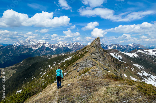 A woman with big hiking backpack hiking to Himmeleck peak, Austrian Alps. There is a massive mountain range in the back, partially covered with snow. Early spring vibes. Freedom and adventure.