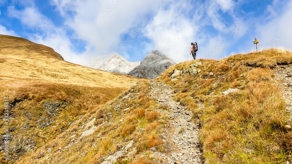 A man with a big backpack hiking through golden colored mountains on the Austrian-Italian border. The man is enjoying the autumn vibes in Alps. Sharp Alpine peaks in the back. Serenity and solitude