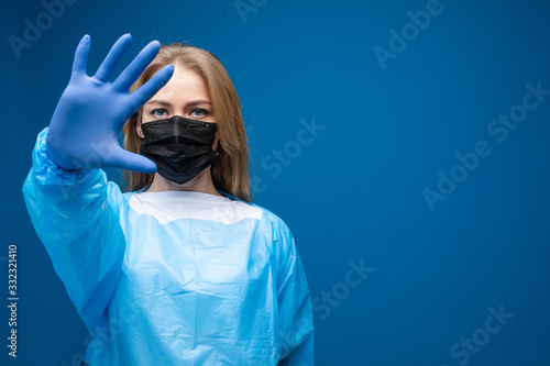 Stock photo portrait of a Caucasian adult woman in black respiratory mask, blue robe and rubber glove showing stop gesture at camera with her hand. Stop coronavirus concept. Copy space. photo