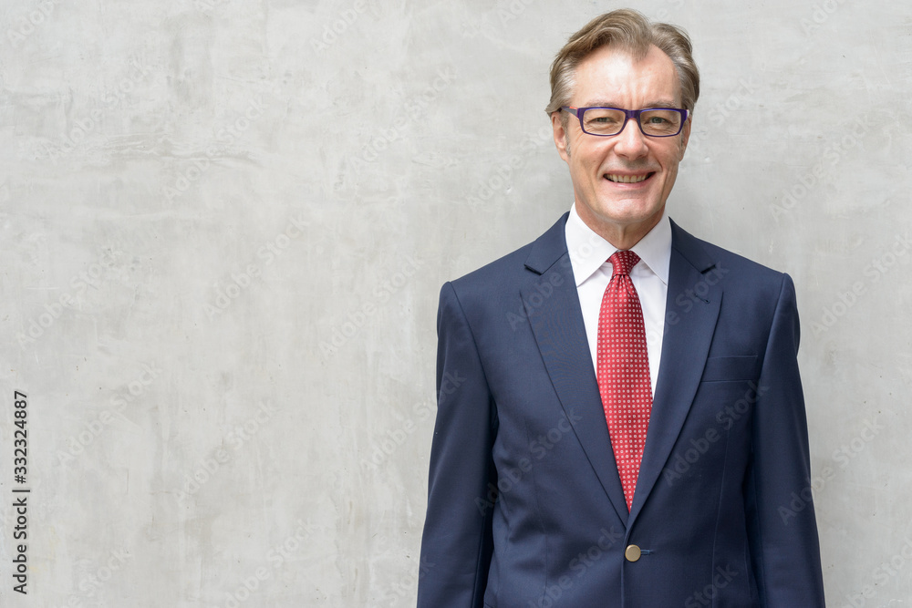 Happy handsome mature businessman in suit smiling against concrete wall Happy handsome mature businessman in suit smiling against concrete wall