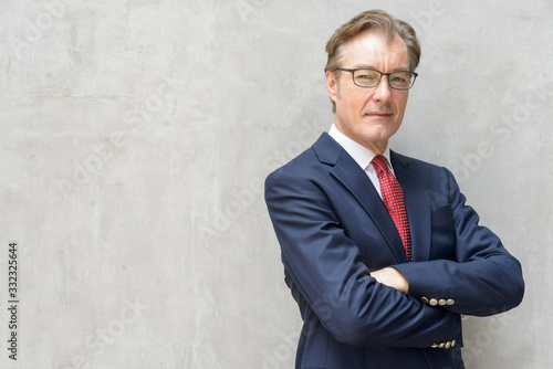 Photos Handsome mature businessman in suit with arms crossed against concrete wall